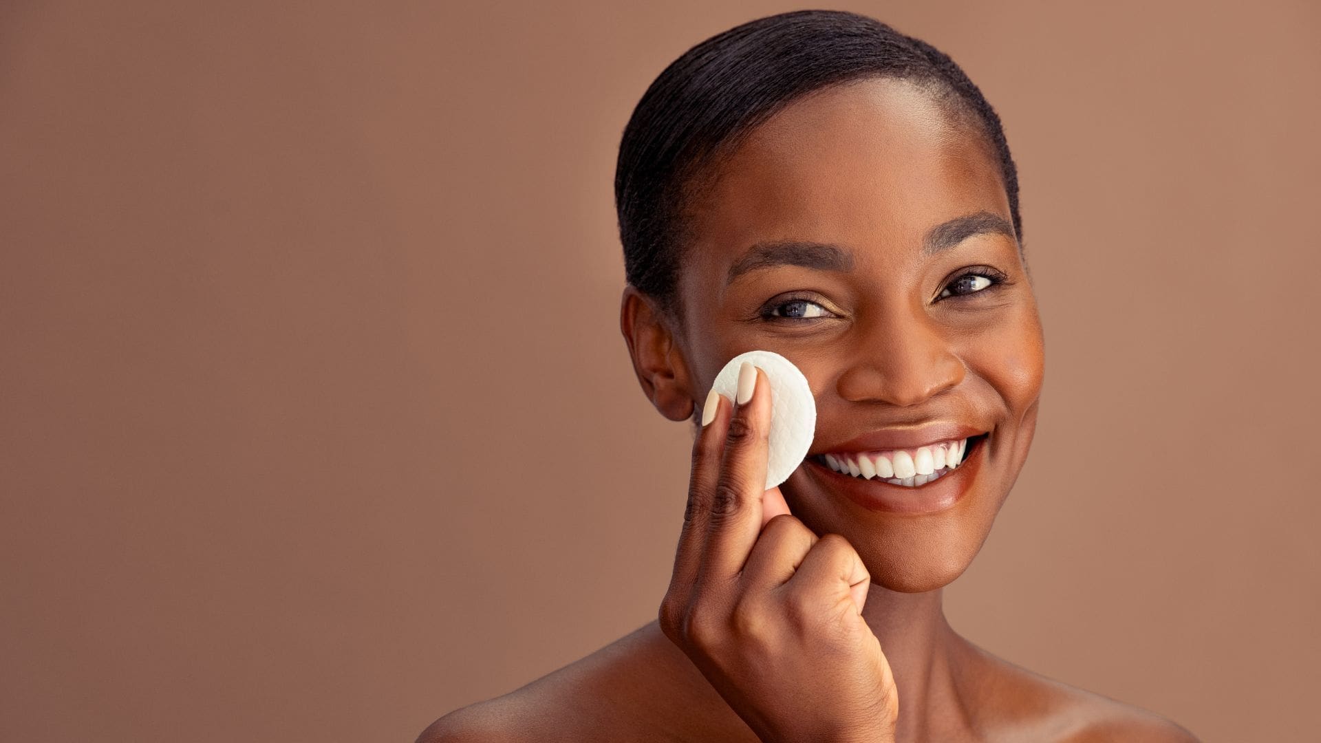Smiling woman using a cotton pad for skincare at Towson Med Spa, showcasing healthy, radiant skin in a neutral backdrop.