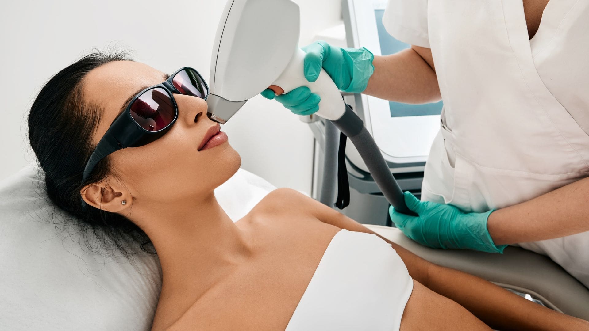 Woman receiving acne treatment facial with LED light therapy in med spa, wearing protective glasses, while technician operates device.