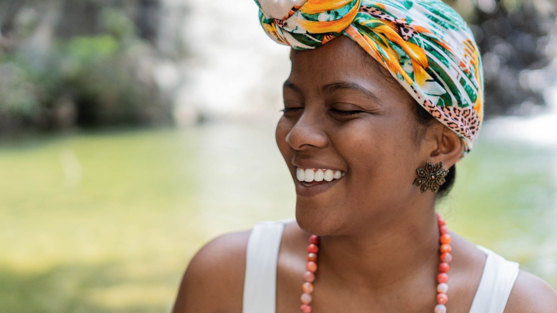 Smiling woman with a colorful headwrap and beaded necklace, enjoying a natural outdoor setting, representing youthful vitality and beauty.