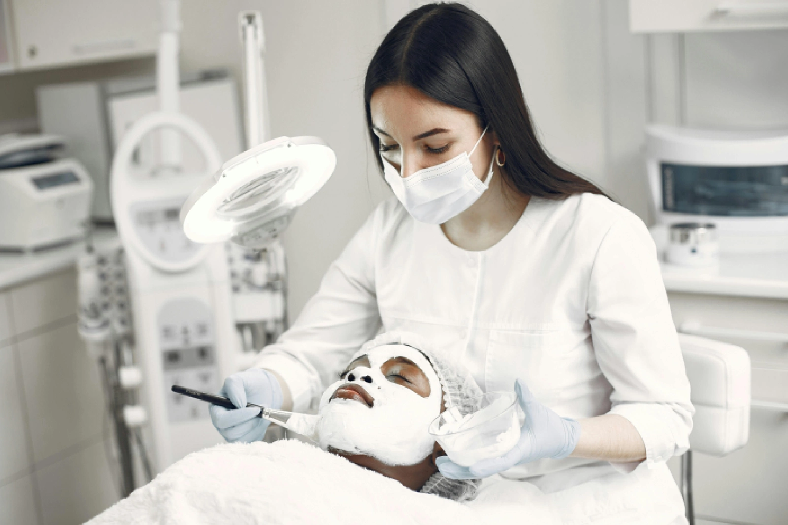 A woman receiving a facial treatment from a masked esthetician.
